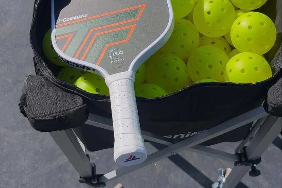 A pickleball paddle resting on a cart full of yellow pickleballs on an outdoor court